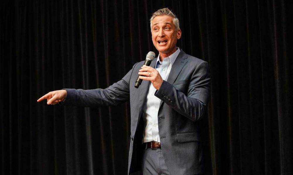 Scott Greenberg speaking into a microphone and pointing to his right while on stage with a black backdrop.