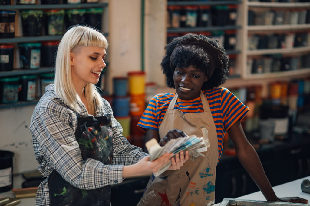 Portrait of multicultural graphic industry female workers standing at printing workshop with color swatch in hands and choosing right color. Interracial graphic technicians choosing colors at workshop