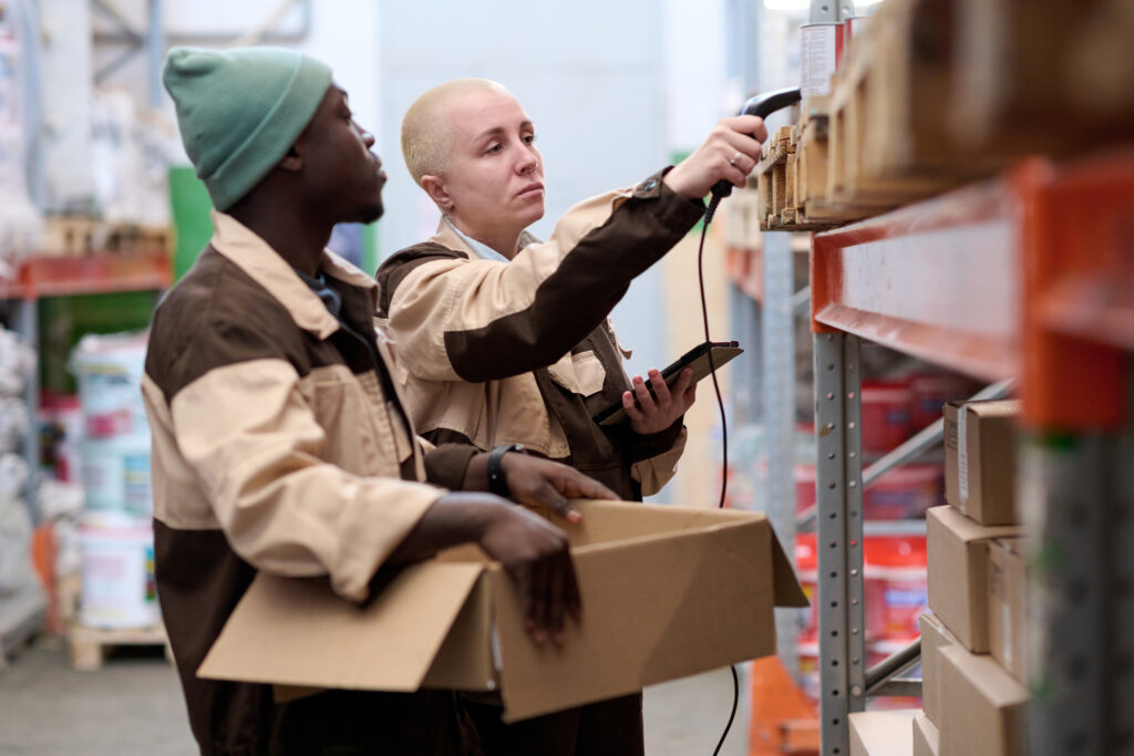 Woman checking barcodes of parcels on shelves with scanner together with her colleague helping her at warehouse