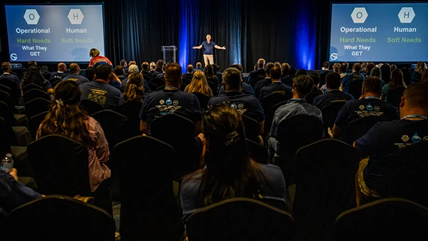 large crowd viewing viewing a presenter on a large stage with arms open wide.